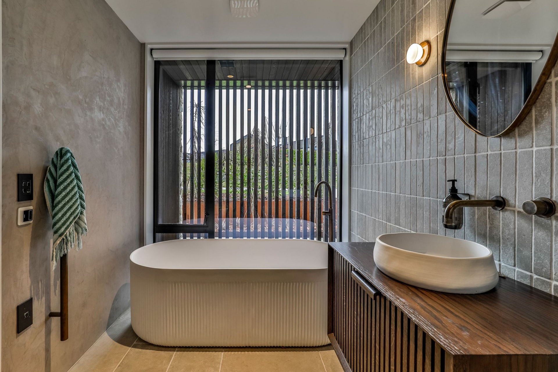 Freestanding fluted bath in front of a full-height window screened by dark battens