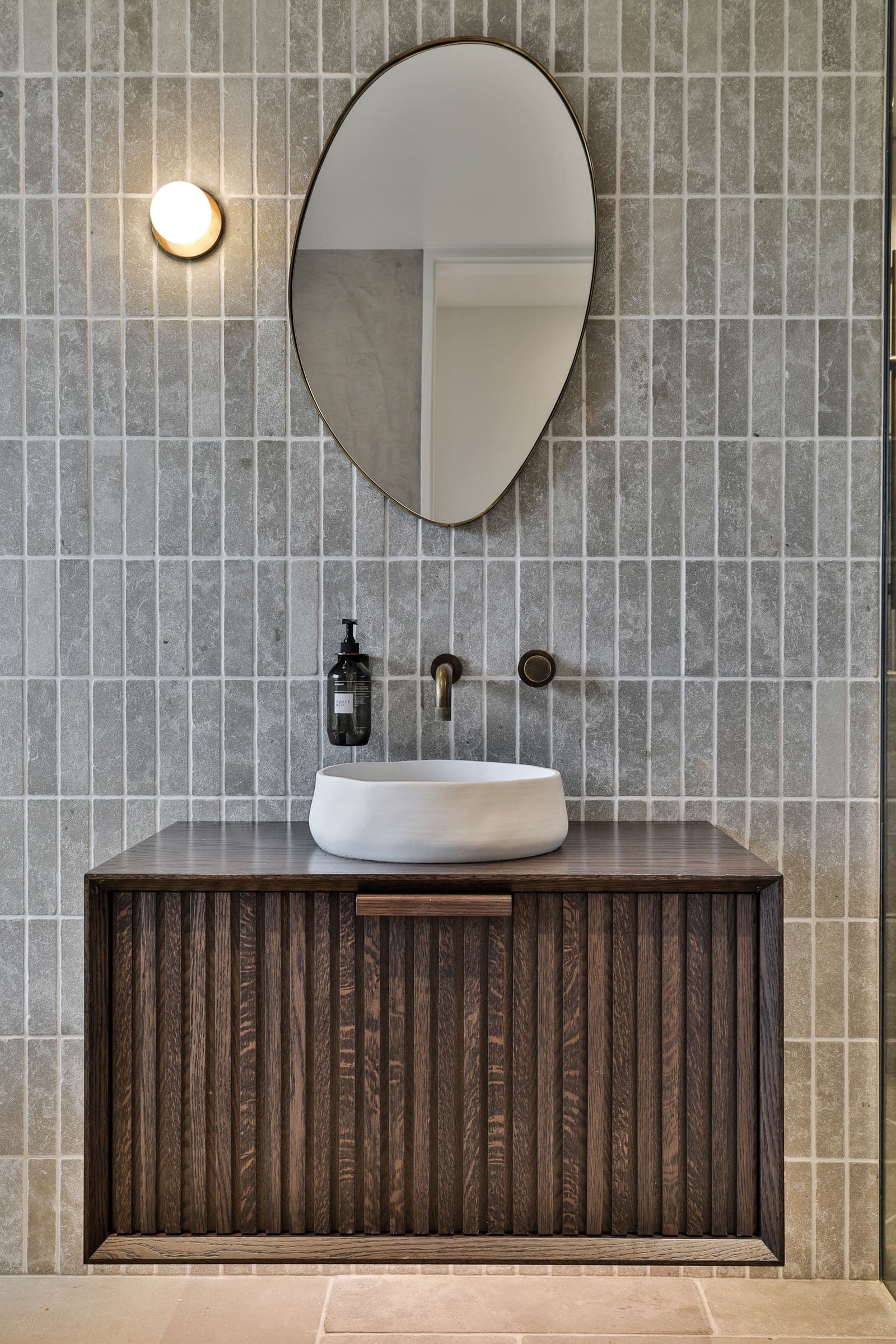 Single fluted oak vanity under a pebble-shaped brass mirror on grey stone finger tile